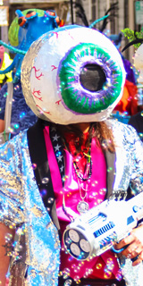 A man in a large eyeball mask and silver sequin jacket.
